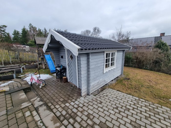 Grå lekstuga, stenlagd väg, gräsmatta, barnets rosa cykel, molnig himmel, uteplats, vit fönsterkarm.