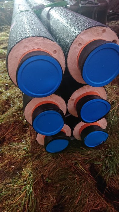 Staplade isolerade rör med blå ändkapslar, vått gräs, utomhus, möjligen byggnads- eller VVS-material.