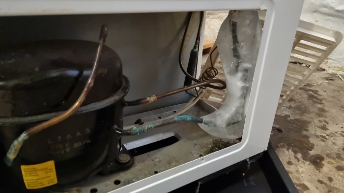 Öppet kylskåp visar kompressor och kondensorrör, med synlig isbildning. Oorganiserad, smutsig bakgrund. Reparationsbehov tycks föreligga.