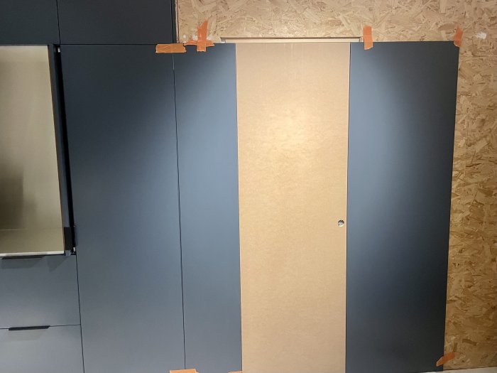 Blåa skåpdörrar monterade på vägg, tejpade kanter, reflektion och oslipad plywood i bakgrunden.