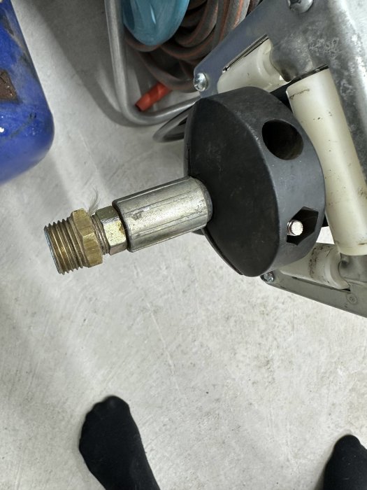 Närbild av en apparat med gängad metallanslutning, svart plasthölje, på ett golv med synliga fötter.