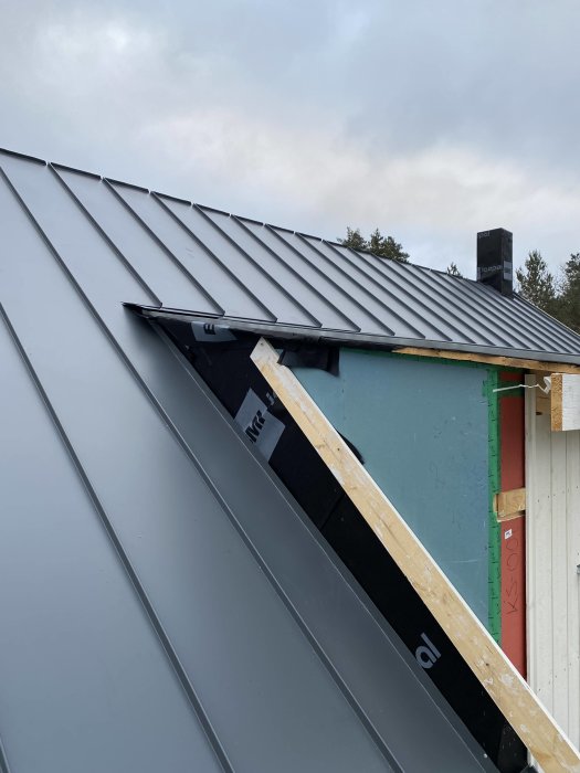 Ett hus under konstruktion med nytt metalltak, taksteg och en skorsten. Oisolerade väggar synliga. Molnig himmel.