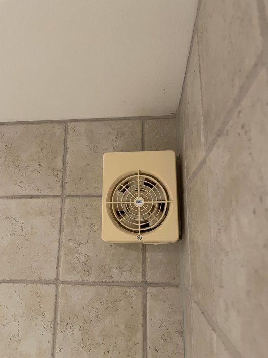 Ventilationsfläkt på vägg, kakel, beige, hörn av rum, lutande perspektiv.