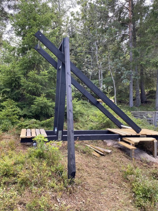 Svart metallkonstruktion i naturen, pågående bygge, trä och verktyg runtomkring, blandad skog i bakgrunden.