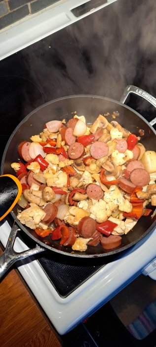 Stekpanna med korv, grönsaker och ånga på spishäll. Matlagning. Lekreusstekpanna. Blomkål, paprika, lök, svampar.