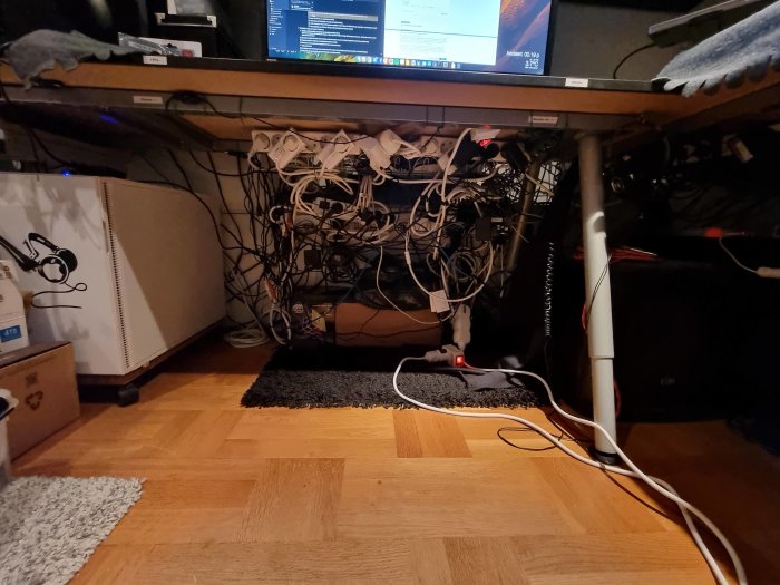 Skrivbord med rörig kablage under, skärmar ovanpå, golv med mattor, kontorsmiljö.