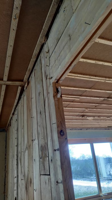 Inomhus träkonstruktion, obehandlad trävägg, spikar, snö synlig genom fönstret, pågående bygg eller renovering.