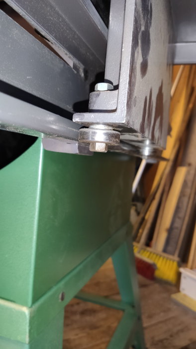 Metallkonstruktion monterad på grön stativ med bult och mutter, oskarp bakgrund med diverse föremål.