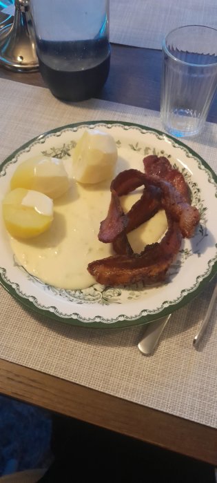 Ett måltidsstilleben med stekt bacon, kokt potatis, sås, ett vattenglas och saltkvarn på bord.