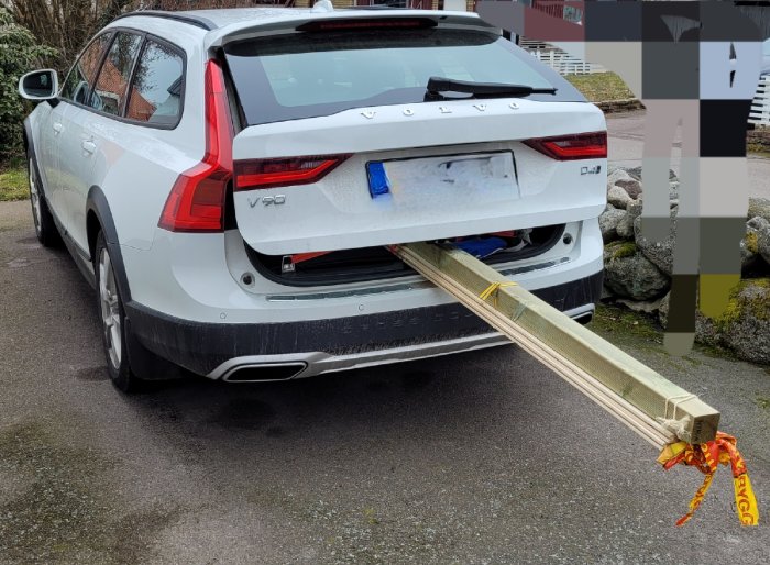 Vit Volvo V90 med långa träplankor som sticker ut genom bakluckan, säkrade med orange spännband.