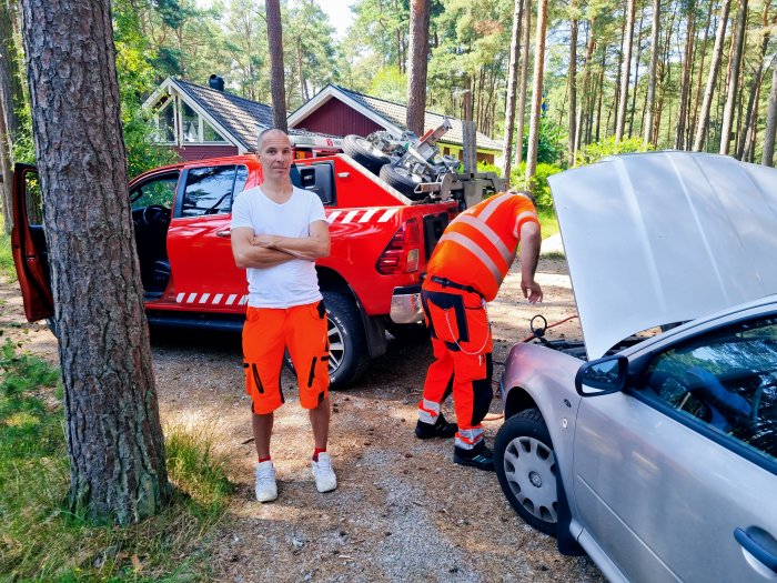 Man i orange står bredvid bil, räddningstjänst på plats, skogsmiljö, bekymrad person framför.