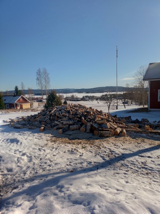 Vinterlandskap med vedhög, snö, röd stuga, flaggstång, skog och blå himmel.