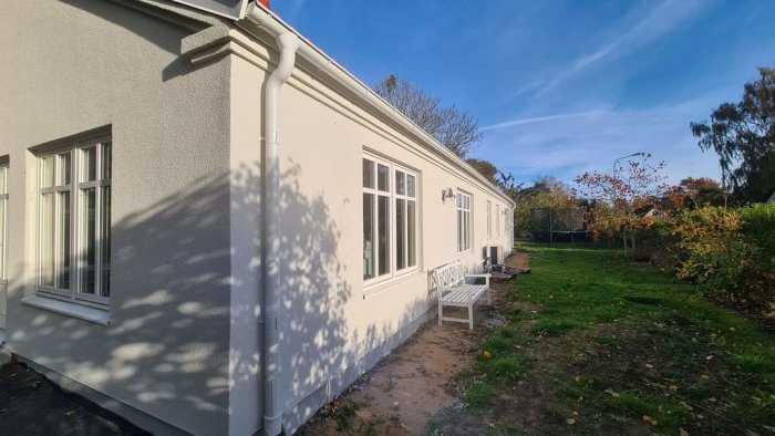 Enfamiljshus med vit fasad i solsken, trädgård, grönt gräs, skuggor på väggen, parkbänk och klarblå himmel.