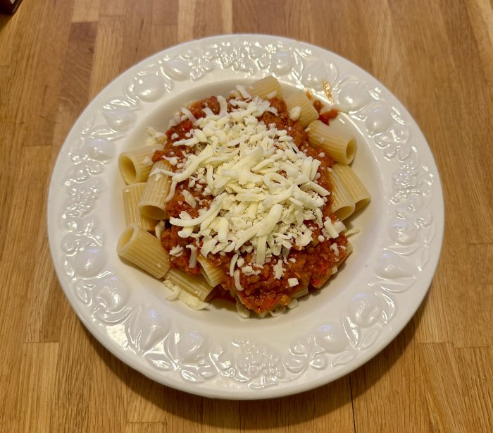 En tallrik med pastarör toppade med köttfärssås och riven ost på ett träbord.