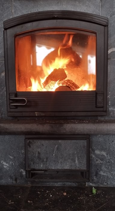 Brinnande ved i en öppen spiskassett, flammor synliga genom glasdörren, på en stenbakgrund.