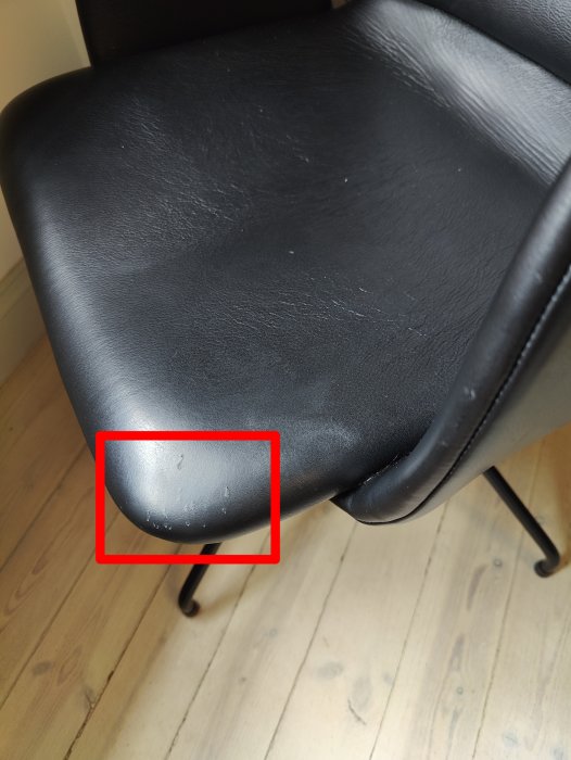 Svart kontorsstol, närbild, synligt slitage på framkant, trägolv bakgrund, röd rektangel markerar skadan.