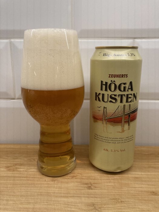 Ölburk märkt "Höga Kusten", glas med skummande öl, neutral bakgrund, träyta, svensk alkoholhalt 5,3%.