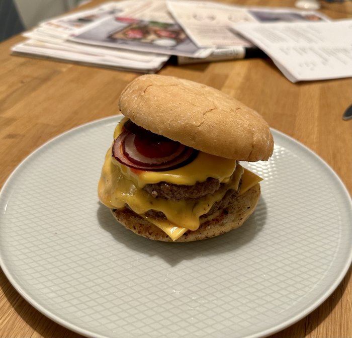 Cheeseburgare med smält ost, lökringar och bröd på tallrik, tidningar i bakgrunden på träbord.