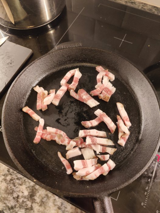 Stekpanna på spishäll med rå baconbitar redo för tillagning, köksmiljö.