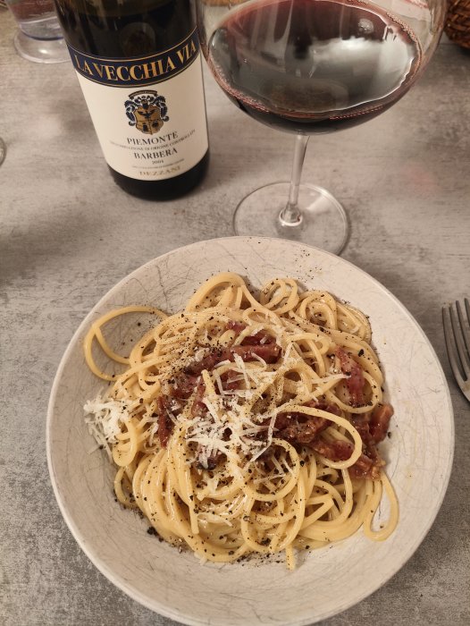 En tallrik pasta carbonara med riven ost, svartpeppar och ett glas rött vin.