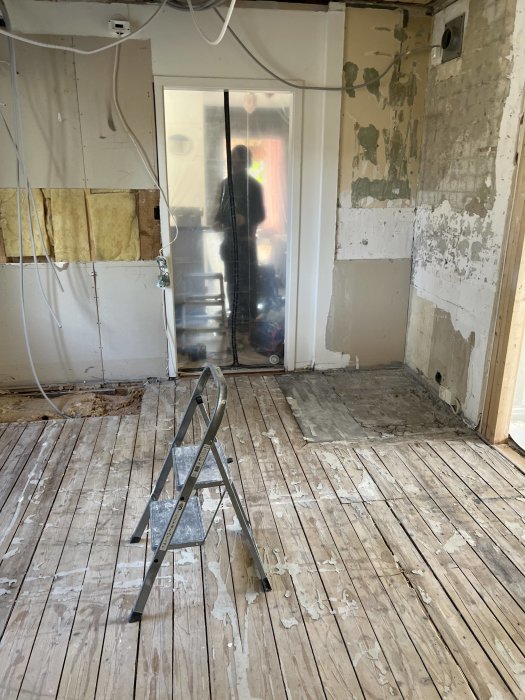 Ett rum under renovering med avskalade väggar, en stege, slitna trägolv och avskavd färg.