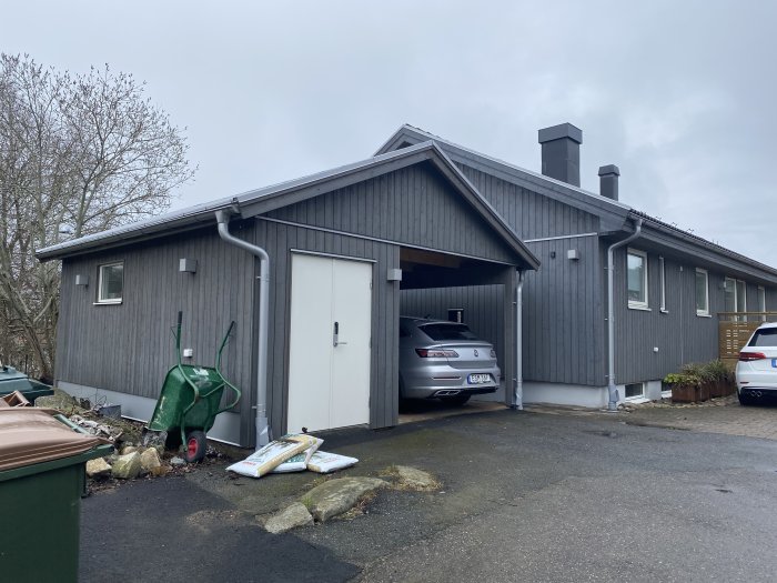 Grått enfamiljshus med carport, bilar parkerade, skräpvagn och sophinkar, molnig dag.