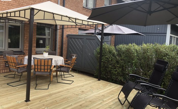 Trädäck med pergola, matplats, loungestolar och parasoll vid tegelhus, grönska, molnig himmel.