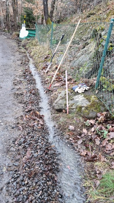 Vattendike vid grusväg, redskap mot staket, våt väderlek, blöta löv, skyfflar, kratta, sten, mossa, arbetsplats.