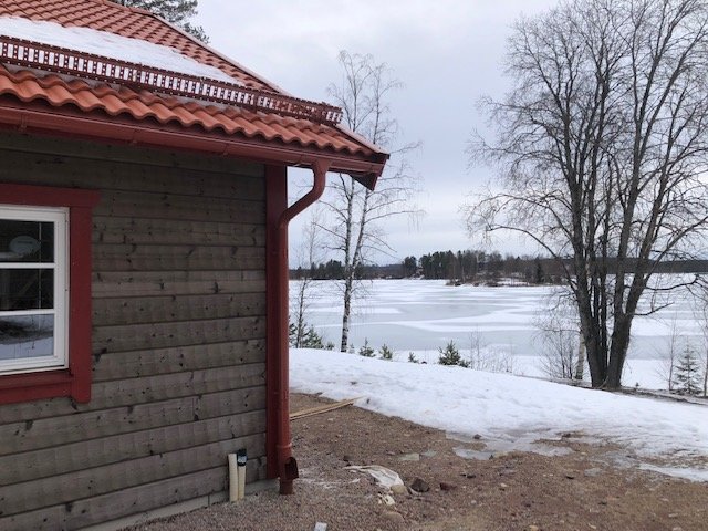 Trähus vid frusen sjö, snöfläckar, barmark, molnig himmel, vinter eller tidig vår.