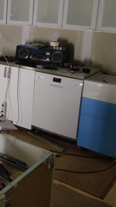 Ett rörigt utrymme med köksskåp, en gammal boombox och en diskmaskin under installation eller reparation.