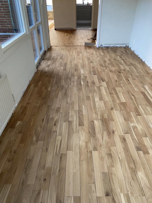 Tomt rum med trägolv, fönster och öppning till annat rum med köksskåp.