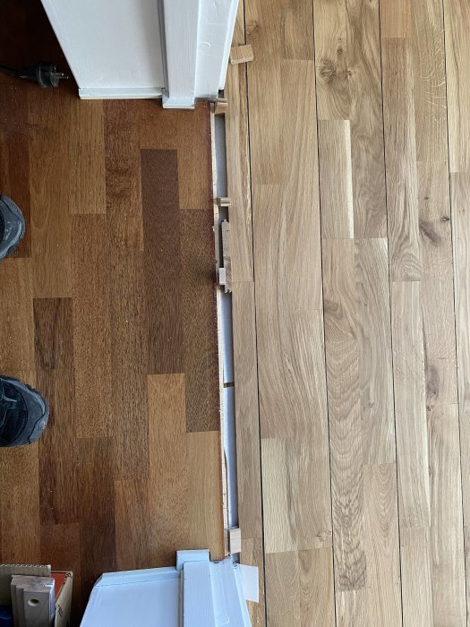 Två olika typer av trägolv möts, möjligen under renovering, med synliga dörrkarmar och personens skor.