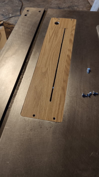 Träplatta med slits och hål, metallunderlag, fyra blå skruvar. Verkstadsmiljö. Arbete eller byggprojekt pågår.