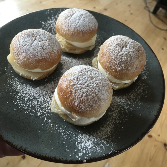 Semlor på mörk tallrik, pudrade med florsocker. Fyra bakverk, fyllning synlig, traditionellt svenskt fika.