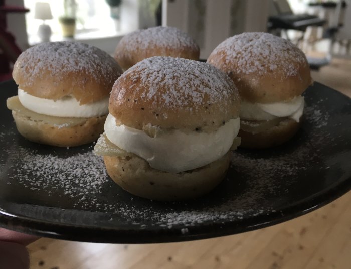 Fyra semlor med grädde och mandelmassa på svart tallrik, pudrade med florsocker.