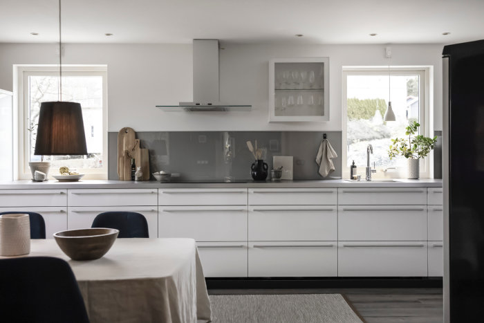 Modernt kök med vita skåp, grå bänkskiva, fönster och hängande lampa.