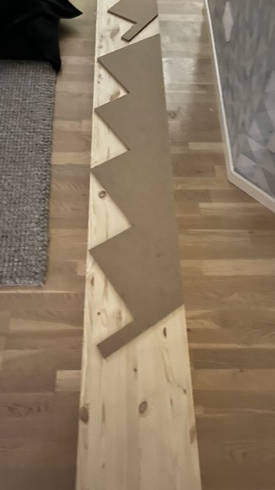 Långt träbräde på golv, grå matta, grått överkast, och bitar kartong ordnade i mönster.
