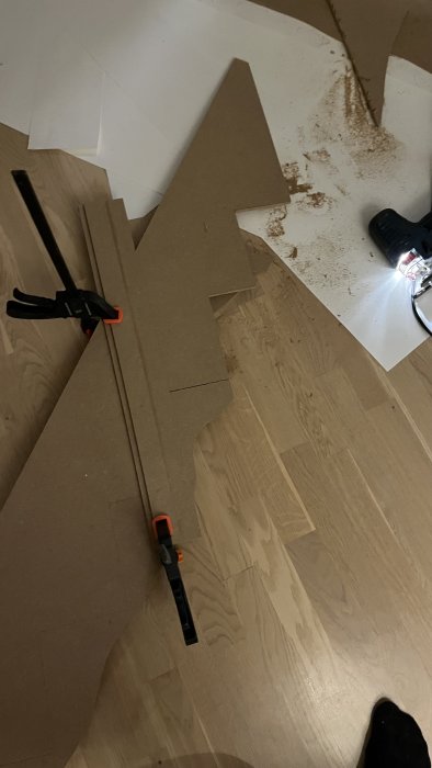 Hantverksprojekt med wellpapp, spill, verktyg och klämma på ett trägolv.