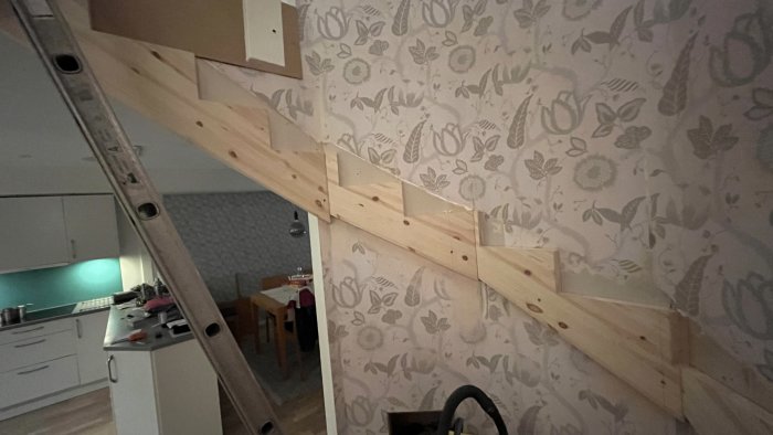Trästege lutad mot blommig tapetvägg; hemmiljö med kök och matplats i bakgrunden.