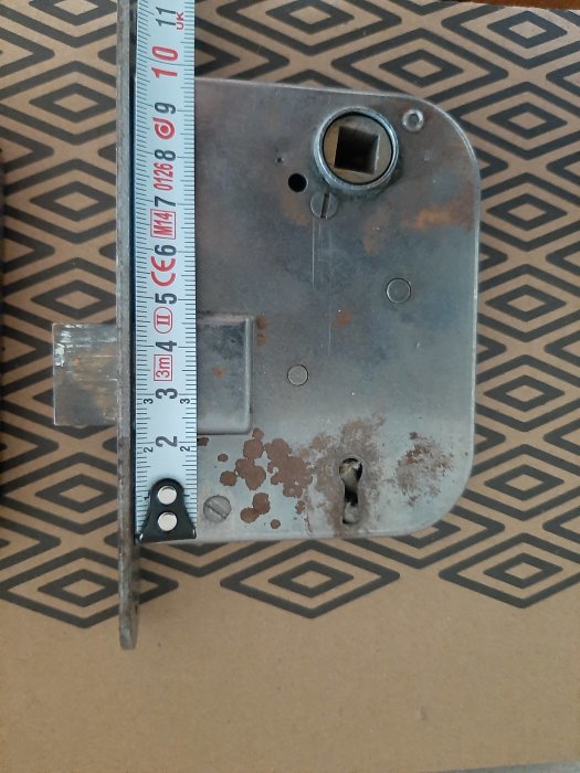 Metallplatta med rost, hål, mäts med tumstock, geometriskt mönstrad bakgrund.