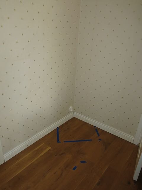 Ett rumshörn med tapet och trägolv, markerat med blått tejp på golvet.