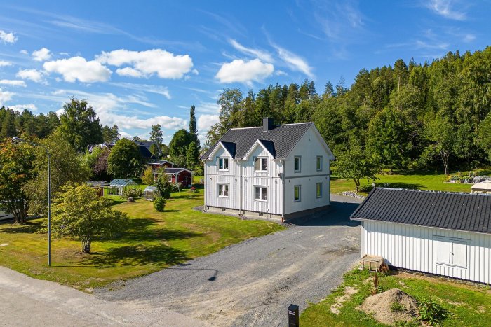 Ljust hus med vita knutar, grönskande trädgård, klarblå himmel med vita moln, lantlig miljö, garage, solsken.