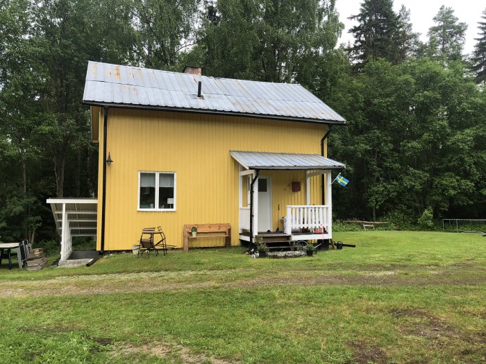 Gult hus med vita knutar, veranda, svensk flagga, omgivet av grönska och gräsmatta.
