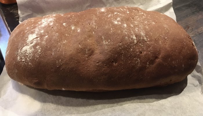 Ett nybakat bröd på bakplåtspapper, mörkt och strösslat med mjöl ovanpå.