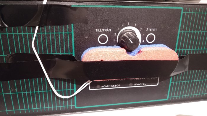 Panel med ratt, tejp, text på svenska, knappar märkta "TILL/FRÅN", "ÅTERST.", "KOMPRESSOR", "GIVARFEL". Green grid background.