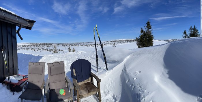 Solig snöig landskap, skidor lutade mot stuga, avkopplande stolar, blå himmel, vildmark.