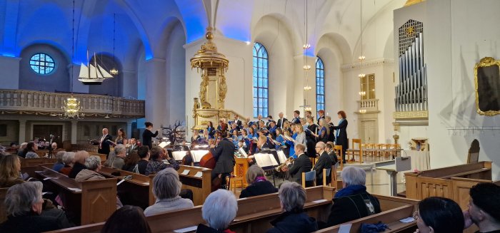 Kör och orkester framträder i kyrka, med åhörare och dirigent, ljus interiör, välvd takkonstruktion, klassisk arkitektur.