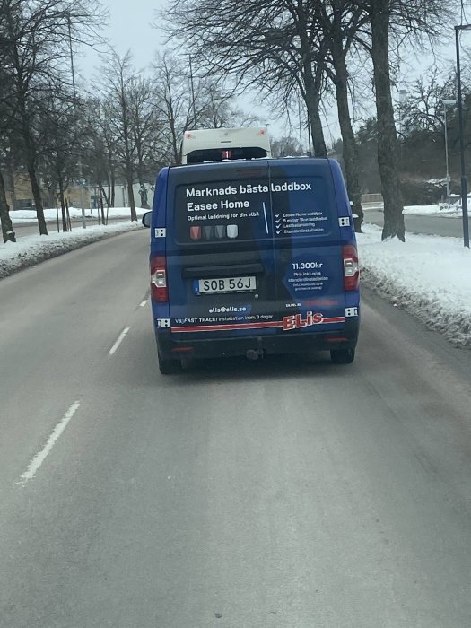 Blå skåpbil med reklam för laddboxar på vinterväg, bar träd, snökanter, registreringsskylt synlig.