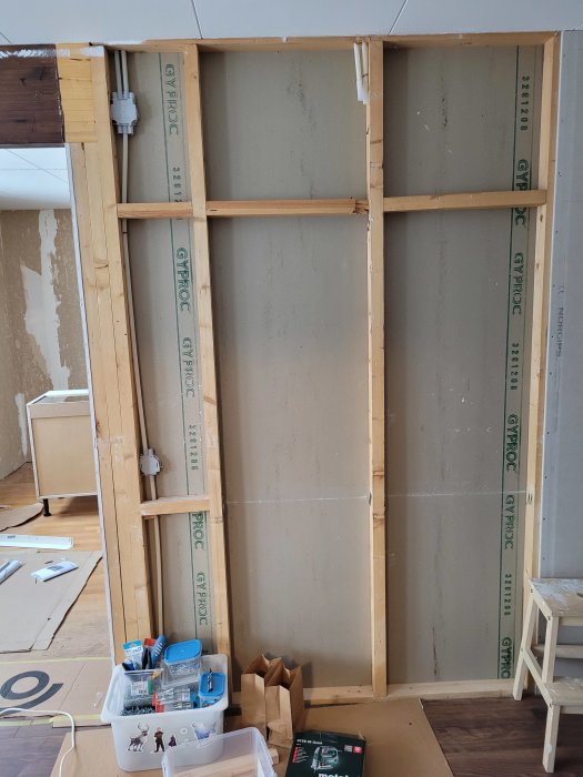 Seinä, jossa puurunko, kipsilevyä ja trippelipaalut vasemmalla. Oikealla kaksi erillistä pystypaalua. Lattialla muovilaatikko ja paperipusseja.