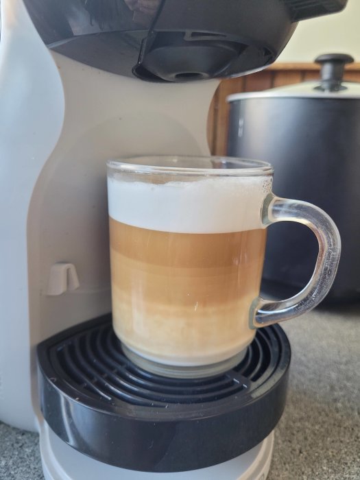 Kaffebryggare laddar en genomskinlig kopp med lager av kaffe och skum, köksmiljö.